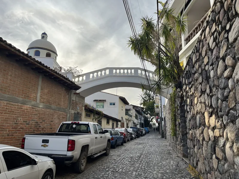 Gepflasterte Altstadtgasse in Puerto Vallarta mit weißer Bogenbrücke über der Straße und einer Kirchenkuppel im Hintergrund