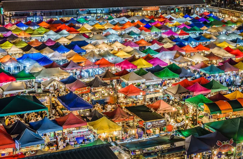 Blick von oben auf die bunten Zeltdächer und Essensstände des Train Night Market Ratchada (Talad Rot Fai) in Bangkok am Abend